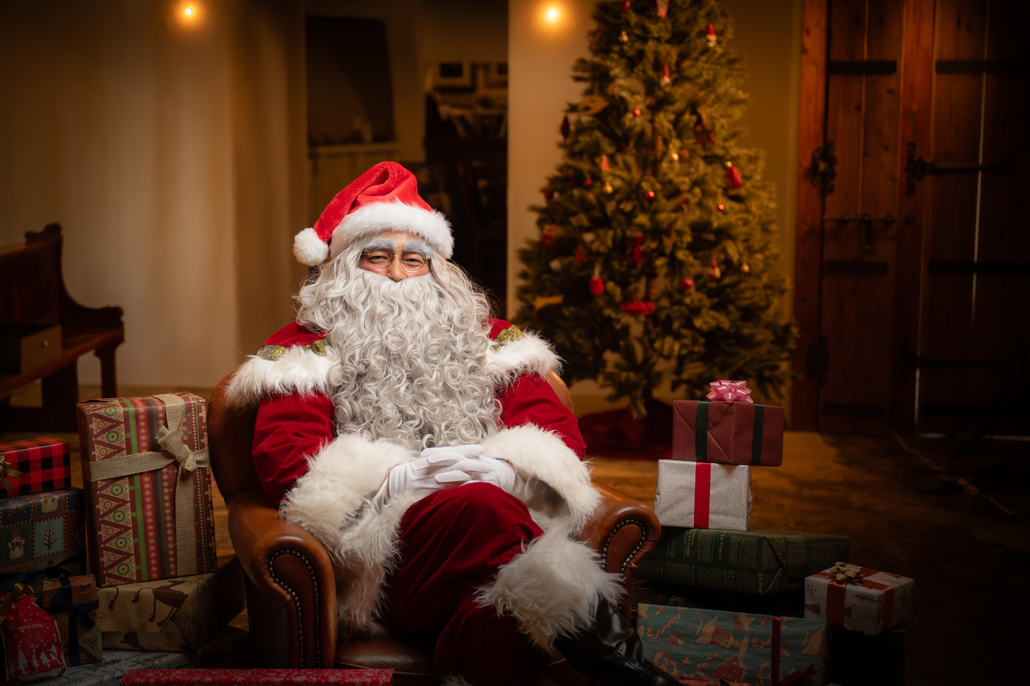 たくさんのプレゼントに囲まれ、クリスマスツリーの前の椅子に座るリアルサンタクロース（サンタ便）